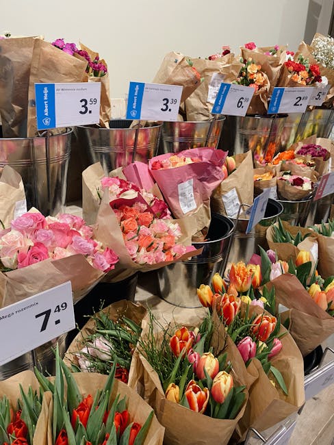 An arrangement of fresh flowers in a florist shop display, featuring pink roses, orange and pink tulips, and mixed bouquets wrapped in brown craft paper. The flowers are organized in metal buckets wit