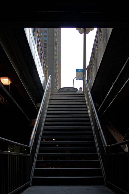 This image shows a metal staircase leading upwards, with a high-rise building visible in the background through an opening at the top. The staircase is flanked by black railings on both sides, and the
