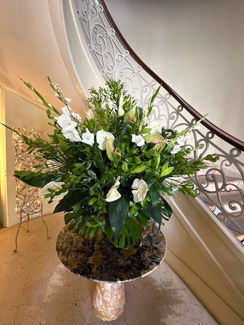 A tall floral arrangement featuring a mix of white flowers, including large calla lilies, anthuriums, and smaller blossoms, complemented by lush green foliage with broad leaves and delicate fronds. Th