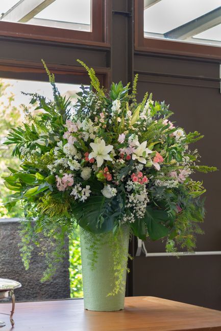 A tall, elegant floral arrangement in a green textured vase, featuring a mix of white lilies, pink and white roses, and delicate smaller flowers, complemented by lush green foliage and fern-like green