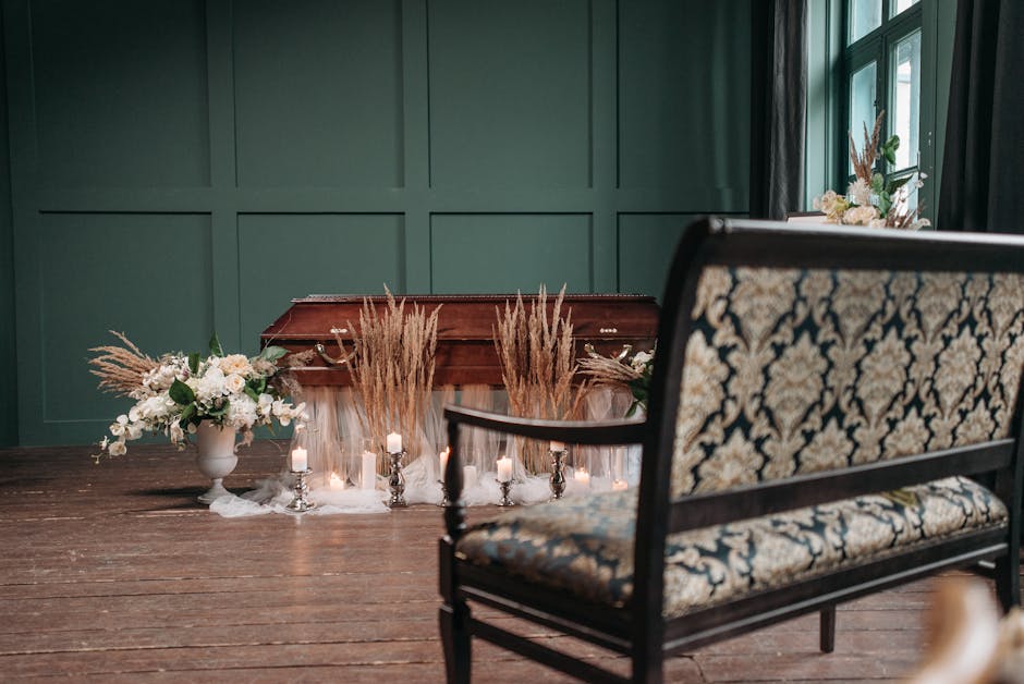 A floral arrangement in front of a closed wooden casket features white and blush pink roses, white lilies, eucalyptus leaves, and dried pampas grass, all artistically arranged in a white urn. Surround