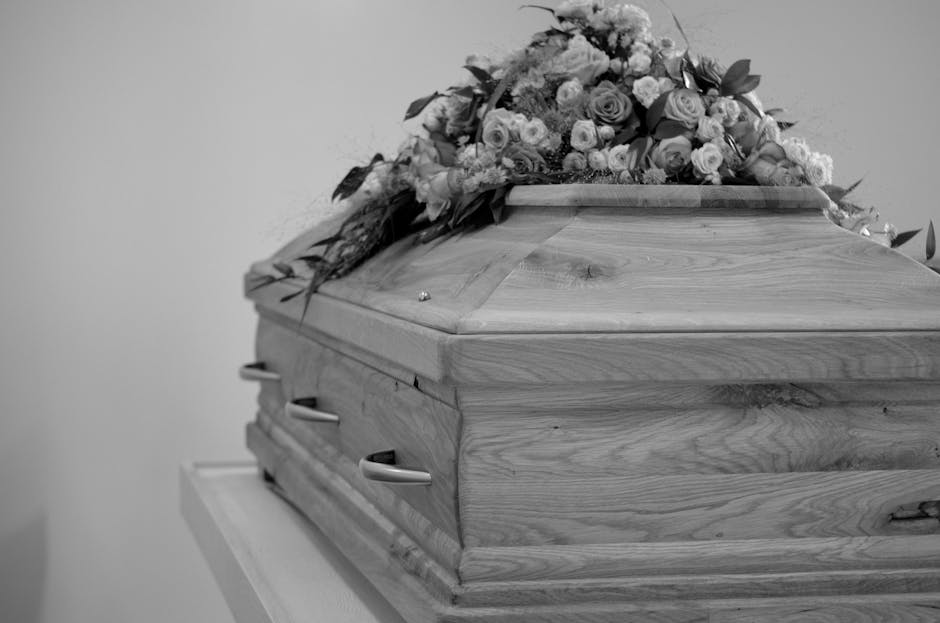 A polished wooden casket resting on a white surface, adorned with a floral arrangement consisting of roses, lilies, and greenery, with soft natural lighting highlighting the textures of the flowers an