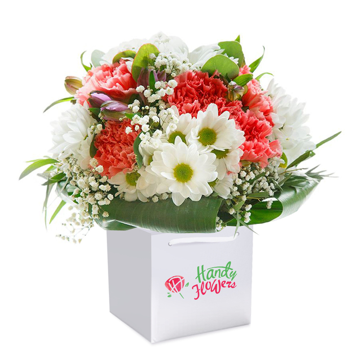 Presented in an immaculate white gift bag, this vibrant arrangement from Cheam Florist radiates a sense of joyful abundance, perfect for brightening homes from the tree-lined avenues of Cheam to the heart of North Cheam. The bouquet is a masterful composition of soft coral and peach carnations, instantly recognizable by their extravagantly frilled petals that create a rich, textured centerpiece. Each carnation is accentuated by clusters of crisp white daisies flaunting lively lime-green centers, bringing a playful contrast and a dash of cheerful charm. Wispy sprigs of baby's breath float between the larger blooms, providing a subtle, romantic softness. Emerging from the left side, a few closed buds with purple-tinged veils add visual intrigue, promising future blossoms. All flowers are encircled by abundant, deep green foliage-broad, waxy leaves that envelop the stems in a natural, supportive cradle while framing the bouquet's rounded profile. The sturdy white ribbon handles atop the square bag reinforce the sense of considered presentation, making the arrangement ready for effortless gifting or delivery. Set against a pristine white background, every detail-from the vibrant flowers to the elegant packaging-is highlighted, ensuring nothing distracts from the bouquet's beauty. Ideal for birthdays, anniversaries, or simply sharing a gesture of appreciation, this local Cheam flower arrangement is a testament to expert design and genuine care, perfect for delivery throughout the area.