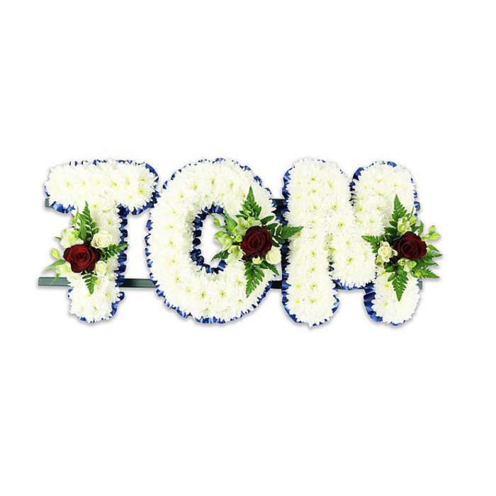 A carefully handcrafted funeral tribute spelling the name TOM in three-dimensional letters composed of countless small white chrysanthemums, each bloom placed to form a dense, cushion-like surface that absorbs light softly and suggests quiet comfort. The letters are crisply defined by a band of royal blue ribbon along their edges, the saturated blue acting like a frame that sharpens the silhouette and adds a refined, respectful contrast against the snow-white petals. Centered within each letter is a restrained floral accent: a single deep red rose anchors the spray, its velvety petals and sombre tone signifying love and remembrance; it is complemented by smaller creamy-white spray roses and tight white buds that create tenderness around the central touch of red. Wisps of fern fronds and glossy green foliage are integrated into the sprays to provide texture, depth, and an organic line that softens the geometric letters. The entire ensemble rests on a discreet dark green or black metal support bar visible along the base, engineered to hold the tribute upright during a service and to ensure the letters remain stable when displayed at a funeral home or church. Photographed against a plain white background under gentle, diffuse lighting, the tribute's colours-pure white, royal blue, deep red and verdant green-read clearly and respectfully. This bespoke memorial piece exemplifies the thoughtful workmanship expected from experienced funeral florists and is intended to convey steadiness, compassion and a personal farewell for family and friends gathered to remember a loved one.