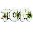 A personalised funeral flower tribute spelling out the name TOM is displayed against a simple white background, each letter crafted as a sturdy, three-dimensional block form secured to a slim, dark support bar. The front of every letter is densely packed with pristine white chrysanthemums, the small, rounded blooms pressed together so closely that they create a smooth, plush surface with a subtle, natural texture. Around the outer edges of the letters, a royal blue satin ribbon has been carefully folded and pinned, forming a crisp, continuous border that sharply defines the shapes and adds a calm, dignified accent. At three key points - on the upper left of the T, the side curve of the O, and the top right of the M - elegant floral clusters add colour and detail. These mini-sprays are centred around deep red roses with velvety petals, symbolising love and remembrance, and are softly framed by smaller white or cream spray roses and slender, white freesia-like flowers. Sprigs of fresh green foliage, including fern and other textured greens, weave through the clusters and gently spill onto the chrysanthemum base, providing contrast and a sense of natural movement. The balanced use of white, blue, and red creates a respectful, quietly striking design that would be fitting for a service in Cheam, whether placed at the front of a chapel, beside a coffin, or by the graveside. It is a classic, lovingly made name arrangement that allows family and friends to honour Tom with a clear, visible tribute during a time of grief and reflection.