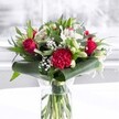A rich, romantic floral bouquet stands proudly in a clear cylindrical glass vase, its neatly trimmed green stems visible through the fresh water and giving a sense of care and craftsmanship. Deep red roses, their velvety petals just beginning to unfurl, cluster at the heart of the arrangement, joined by a large crimson carnation whose ruffled layers create a soft, tactile focal point. Around these passionate reds, creamy white blooms unfold: elegant white alstroemeria with fine speckles at their throats, plump buds of white lilies poised to open in the coming days, and rounded white chrysanthemums that add a cushion of gentle brightness. Wisps of white gypsophila thread between the larger flowers like tiny stars, adding airiness and a feeling of quiet celebration.
Lush greenery cradles the bouquet, from broad, glossy dark green leaves carefully curled and wrapped around the base to more slender, eucalyptus-style stems that stretch upwards, providing contrast in both shape and shade. The clean white surface beneath the vase and the softly blurred, light-filled background suggest a windowsill or table in a bright Cheam home, perhaps catching the soft afternoon light that often spills across nearby parks and leafy streets. The overall mood is one of thoughtful romance and reassuring warmth, as if the flowers have just been placed down to welcome someone home. This is the kind of design a local Cheam florist would recommend for an anniversary dinner, a Mother's Day surprise, or a special birthday, where the bold red and white palette can express heartfelt affection, admiration, and the joy of being remembered.