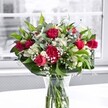 A luxurious bouquet of rich red roses, plush red carnations, and snowy white alstroemeria stands proudly in a clear glass vase on a pristine white tabletop, bathed in soft daylight from a large window. Each rose shows off velvety crimson petals, some fully open to reveal their spiralled hearts, others still in bud, suggesting more beauty to come. Nestled between them, ruffled carnations echo the same deep red tone, adding volume and a classic, almost nostalgic charm. Clusters of white alstroemeria, a mix of tight buds and partially opened trumpets tipped with fresh green, thread through the arrangement, their creamy petals lifting the overall design and softening the intense reds. Tiny stars of white gypsophila, or baby's breath, drift delicately through the bouquet like a fine mist, creating a gentle, cloud-like halo. Around the outside, broad, glossy green leaves and lighter, more slender foliage form a lush collar, framing the flowers with sculpted texture and giving the piece a sense of structure and movement. The stems, neatly cut and visible through the water in the vase, underline the freshness and care of the design, much like a bouquet freshly delivered to a home off Cheam High Street or near Nonsuch Park. Behind, the view through the window is softly blurred, rendered in muted, almost greyscale tones so that nothing distracts from the vibrant reds, crisp whites, and layered greens. The overall mood is celebratory yet refined, perfect for a romantic anniversary dinner, Mother's Day surprise, or a thoughtful thinking of you gesture that brings a touch of elegant joy into any Cheam living room.