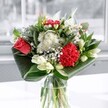 A clear glass vase holds a thoughtfully styled bouquet that feels like a cool breath of Cheam morning air, as if set on a windowsill overlooking the green calm of Nonsuch Park. At the heart of the arrangement, a single coral-red rose and a rich crimson carnation draw the eye, their velvety petals layered and plush, full of quiet intensity. Around them, clusters of tiny white, daisy-like blossoms and small button chrysanthemums create a soft, clouded halo, their petals crisp and clean against the deeper hues. Creamy white lilies appear mostly in bud, tall and elegant, their closed forms promising to unfurl in the coming days and add another chapter of beauty to the display. Broad, glossy emerald leaves are folded and curved at the base, wrapping the stems in graceful arcs that frame the flowers and lend the design a distinctly modern, sculptural silhouette. The natural daylight streaming from a nearby window bathes the bouquet in a cool, even brightness, catching the sheen on each leaf and illuminating the subtle textures in the petals. There is a sense of balance and stillness here, as if the flowers have been arranged to pause time for a moment, perfect for celebrating a new home near Cheam Village, to mark a birthday with understated elegance, or simply to bring freshness and uplift into a quiet corner of the house. You can almost imagine the delicate, green-fresh scent and faint sweetness that would greet you as you lean in, a gentle reminder of the changing seasons outside.