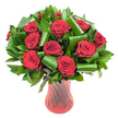 A lush, rounded bouquet of roughly a dozen deep crimson roses sits like a jewel in a slender ruby-red glass vase, the scene captured against a clean white backdrop and soft, flattering light. Each rose is fully open with velvety petals spiralling from a tight heart to a lavish outer bloom, the color leaning toward wine-dark crimson where shadows fall and a more brilliant ruby where the light catches. Surrounding and supporting the rose heads is abundant dark green foliage - glossy, flat leaves that read like Ruscus or Salal and broader, emerald Aspidistra-style leaves artfully folded, rolled, and curled into sculptural loops. Those green curls are folded with confidence, creating elegant, architectural arcs that cradle the flowers and add modern structure to the classic composition. The vase is translucent, ruby-hued glass with a subtle ribbed or twisted texture that creates vertical lines of reflected light, echoing the roses' rich tones and giving the whole arrangement a slightly dramatic yet refined silhouette. Imagine the soft, classic scent of roses rising from the clustered stems, a warm, familiar perfume that suits anniversaries, Valentine's gestures, or a refined table centrepiece. The white background isolates the arrangement so the reds and greens sing together - perfect for editorial images about romantic gifts, luxe floral design, or celebrations in and around Cheam Village and Nonsuch Park. As a local florist might advise, the design balances old-world passion with contemporary leafwork to feel both timeless and unmistakably hand-crafted in Cheam.