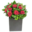 In this elegantly modern composition, rich crimson roses take centre stage, each bloom opening in velvety spirals that catch the light like softly glossed fabric. The roses are set into a compact cluster and cradled by broad, glossy green leaves that have been carefully curled and layered to create a rhythmic, sculptural movement - a tide of emerald that both supports and contrasts the deep red. The arrangement sits in a matte black square vase, its geometric lines grounding the design with a contemporary, urban edge that would suit a chic Cheam apartment or a smart office near Cheam Station. Lighting in the photograph is clear and even, picking out the sheen on each leaf and the soft sheen of the rose petals so their crimson depth reads almost tactile: you can imagine the silken give of a petal beneath your fingertips. There is a restrained freshness to the greenery - almost tropical in its crispness - that lifts the classic rose fragrance into something lighter and more modern. As a local florist describing this piece, I hear it speaking of purposeful elegance: a gift for anniversaries, a declaration of affection, or a way to bring considered beauty to a dinner table in Cheam Village. The craftsmanship shows in the folds of foliage and the precise clustering of blooms, a deliberate choreography that feels both intimate and confident. Close your eyes and the scent is clear: the heady sweetness of red rose brightened by green leaf notes, a sensory reminder of how simple materials, arranged with care, can create a memorable, emotion-filled statement.